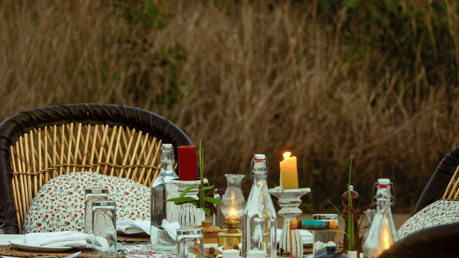A vertical view of an elegantly set outdoor dinner table with bottles, glasses, and wicker chairs against wild grass - Umaria Safari Camp