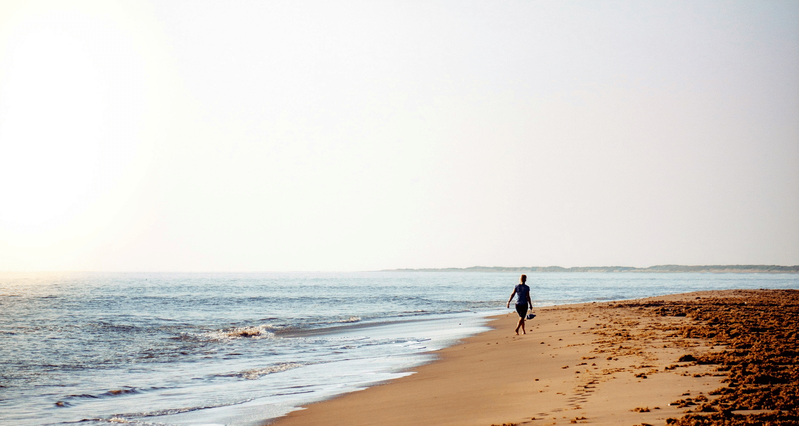 The ocean meeting the sand under a bright sky with a distant figure.