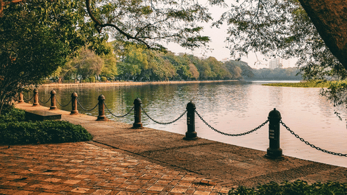 Brick-paved lakeside promenade shaded by mature trees in a landscaped park.