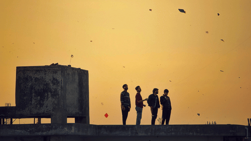 A silhouette of a group of 4 friends standing on a terrace flying kites in the evening.