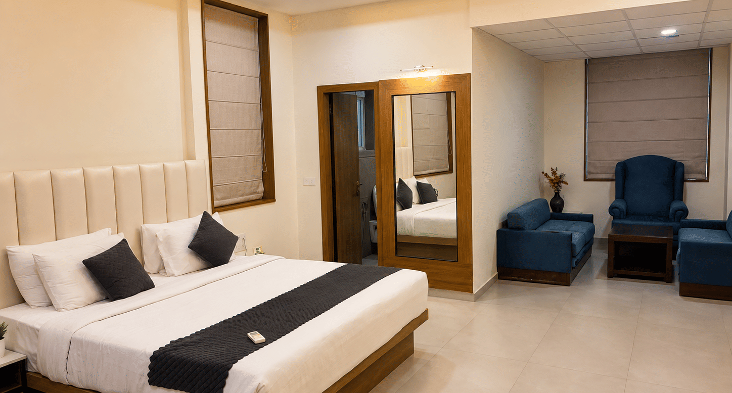 Suite at TGI Apple Inn, Ajmer Road, Jaipur featuring a bed with pillows, a seating area with armchairs and a table, blinds on the windows, and a doorway.
