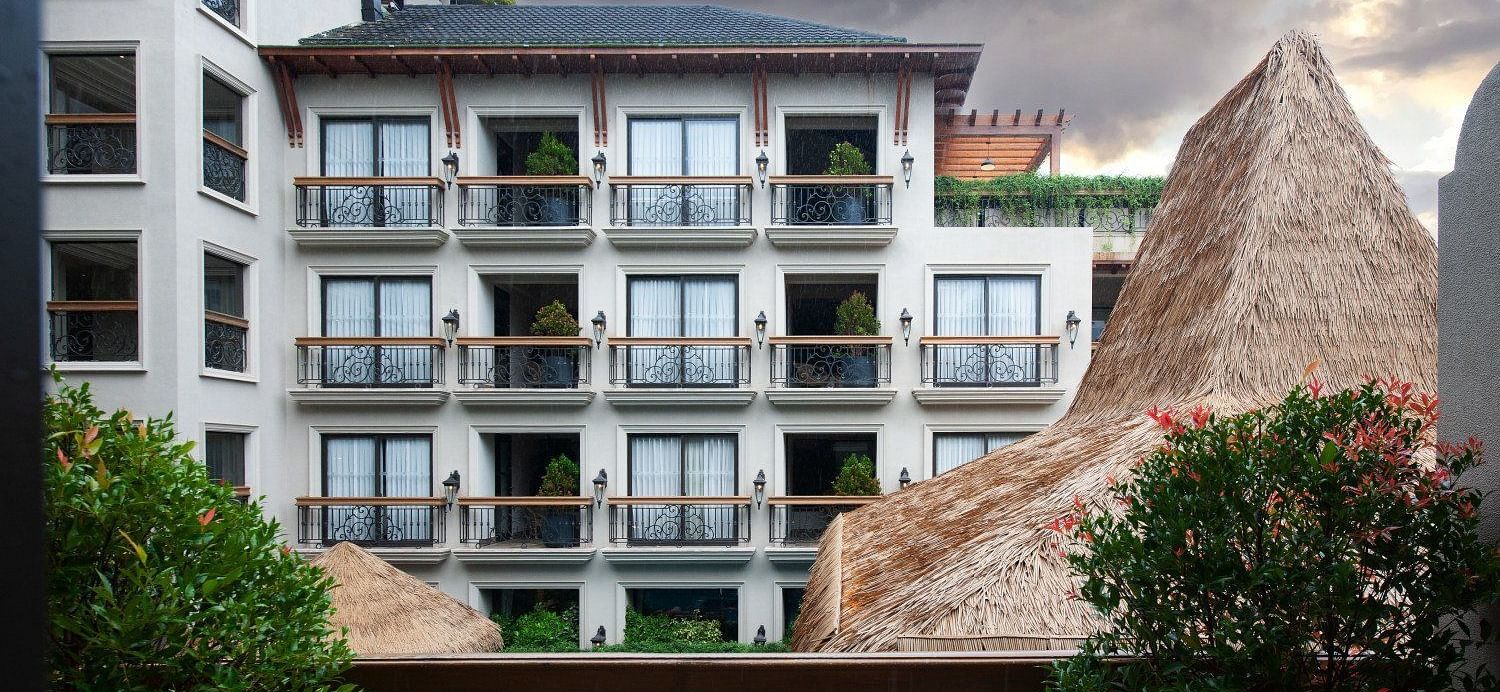 An exterior view of the multi-story hotel building at The Evren, Vagator, with a view of balconies and the swimming pool below. The image shows the building's architecture and the surrounding tropical environment under a dramatic sky.