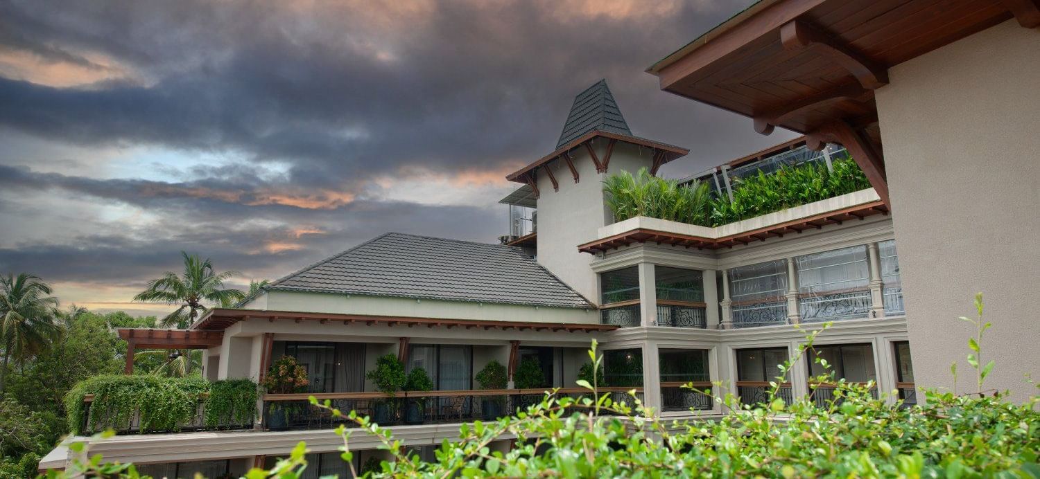 An architectural shot of the exterior of The Evren, Vagator, showing the elegant facade, balconies, and the lush green surroundings. The image is taken from a vantage point that captures the building's design against a dramatic sky.