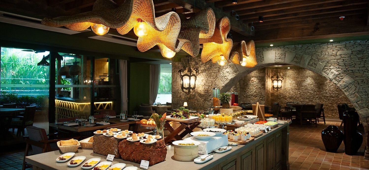 A lavish breakfast buffet spread at Mila Restaurant at The Evren, Vagator, featuring a variety of dishes on a large central island. The restaurant has a rustic yet modern ambiance with woven light fixtures, a stone archway, and a view of the outdoors.