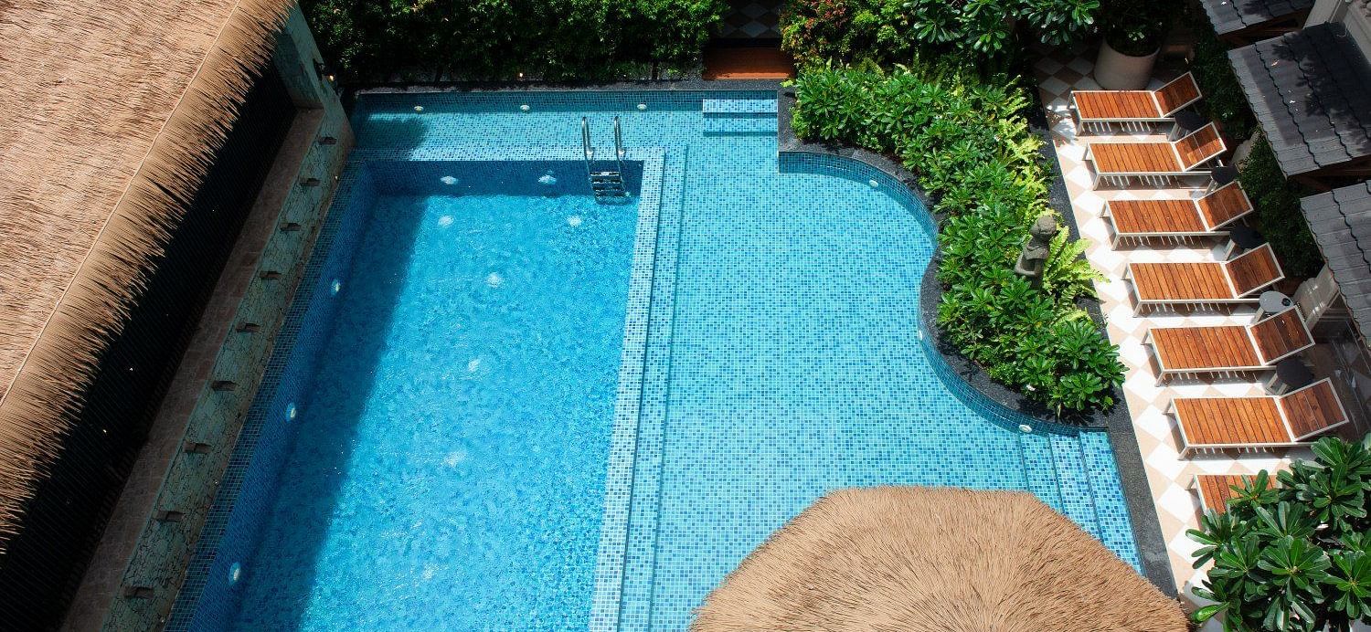 An aerial view of the outdoor swimming pool at The Evren, Vagator, featuring bright blue mosaic tiles, a built-in seating area, and a row of lounge chairs shaded by a large straw umbrella. The pool is surrounded by lush green plants and is overlooked by th