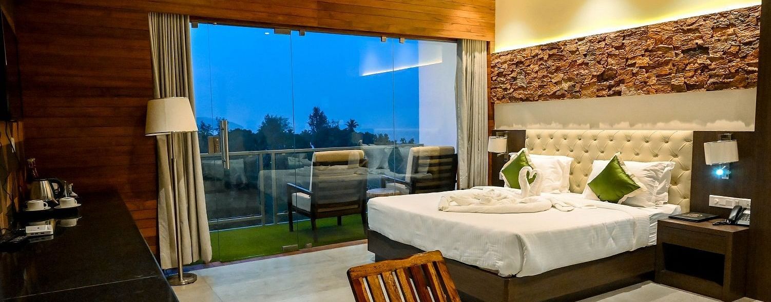A hotel bedroom with a large bed, a comfortable armchair, and a wide view of the outdoors through a large glass door leading to a balcony - Sea Hills Hotels, Sri Vijaya Puram (Port Blair)