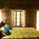 A warm stone cabin bedroom featuring a made bed with bright blue pillows, a patterned yellow bedspread, and natural light streaming through windows.