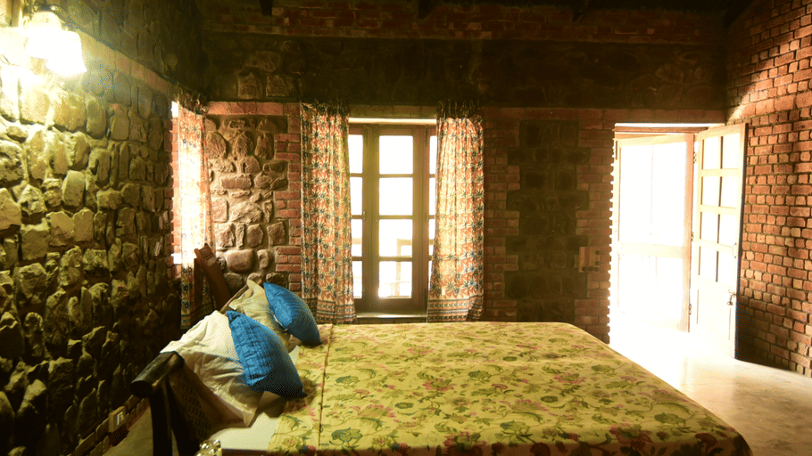 A warm stone cabin bedroom featuring a made bed with bright blue pillows, a patterned yellow bedspread, and natural light streaming through windows.