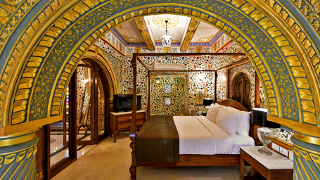A luxurious palace bedroom at Gogunda Palace framed by an ornate gold and blue arch, featuring a wooden 4-poster bed and opulent decor.