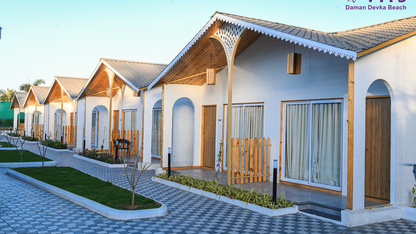 A view of the cottages' facade with cobbled pathway, a small lawn in between the pathway and wooden gates on the entrance to the cottage at VITS Daman Devka Beach.