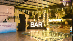 An event venue featuring a bar counter with neatly arranged bottles, glass table setups, and warm lights hanging from the ceiling for decoration | VITS Daman Devka Beach