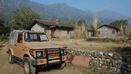 A beige safari jeep parked on a dirt path in front of stone cottages with misty mountains in the background.