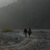 2 people walking along a dusty dirt track in a misty valley surrounded by dark hills.