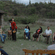 A group of people sitting in a circle outdoors near a thatched shelter and green hills.