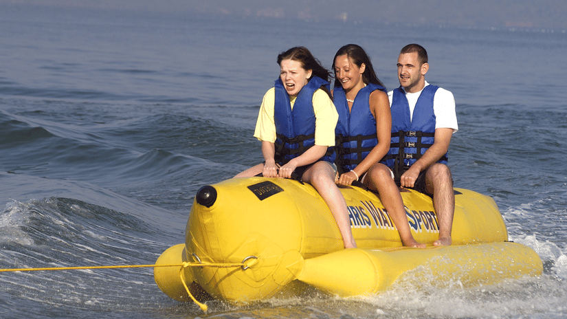 A view of three people sitting on a banana boat while being tugged by another boat.