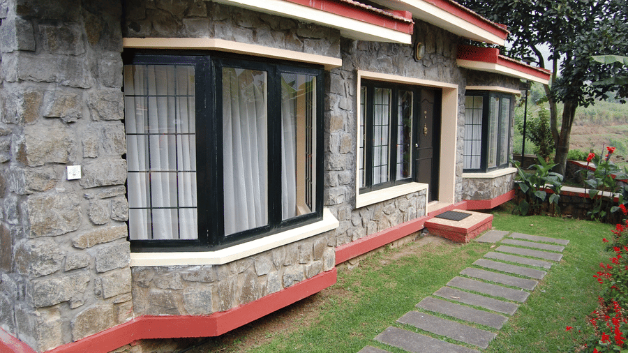 A charming and rustic stone cottage exterior at Green Pastures, Kodai, featuring prominent bay windows and a welcoming flagstone pathway leading across a manicured lawn toward the entrance.
