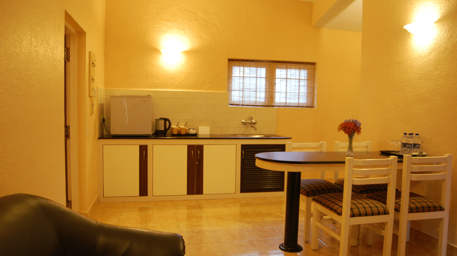 A bright kitchen and a dining area of Deluxe Cottage at Green Pastures, Kodai, featuring clean lower cabinets, a small preparation counter under a window, and a wooden table with seating for four.