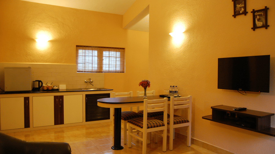 A spacious and fully equipped kitchenette and dining area of Deluxe Cottage at Green Pastures, Kodai, featuring a clean counter space, a compact dining table set, and a modern wall-mounted television.