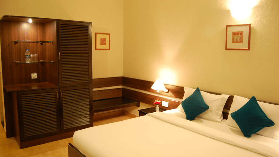 The luxurious Deluxe Room of Green Pastures, Kodai, featuring a beautifully appointed bed with plush pillows and a prominent dark wood built-in wardrobe and refreshment center.