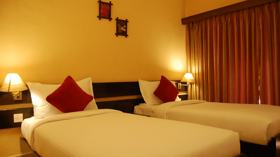 Twin bed with white linens and dark red cushions, bedside tables with lamps, and yellow painted walls in a guest room at Green Pastures, Kodaikanal.