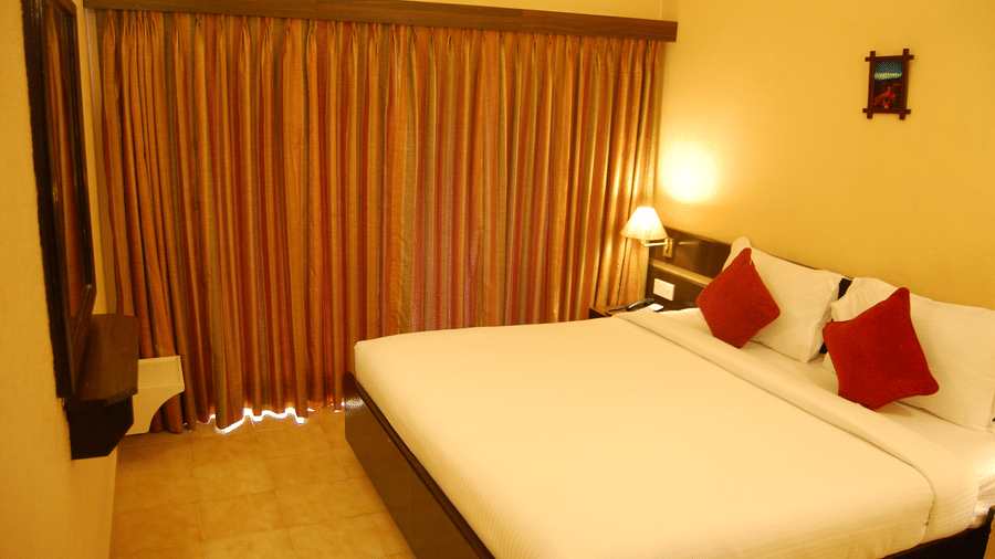 A top corner view of the premium cottage with white bedding and red cushions, curtained window, bedside lamps, and yellow walls in a guest room at Green Pastures, Kodaikanal.