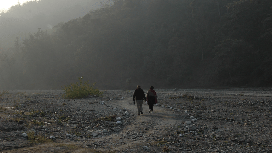 2 people walking along a dusty dirt track in a misty valley surrounded by dark hills.