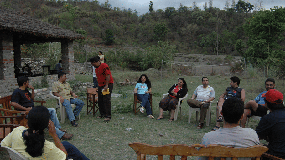 A group of people sitting in a circle outdoors near a thatched shelter and green hills.