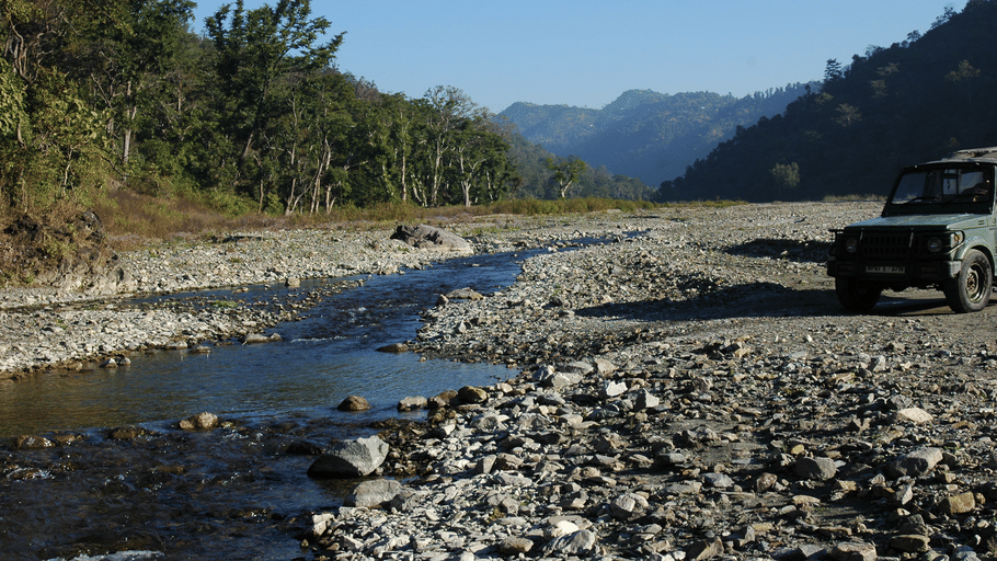 A safari vehicle parked beside a shallow river flowing through a rocky valley surrounded by lush green trees and distant mountains.