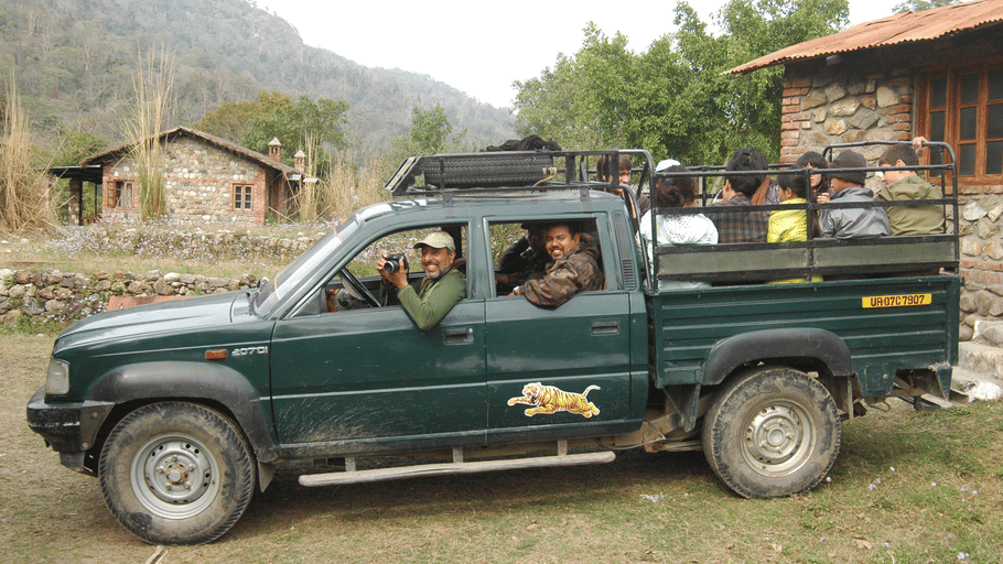 A green safari pickup truck carrying passengers near cottages in a mountainous resort setting.