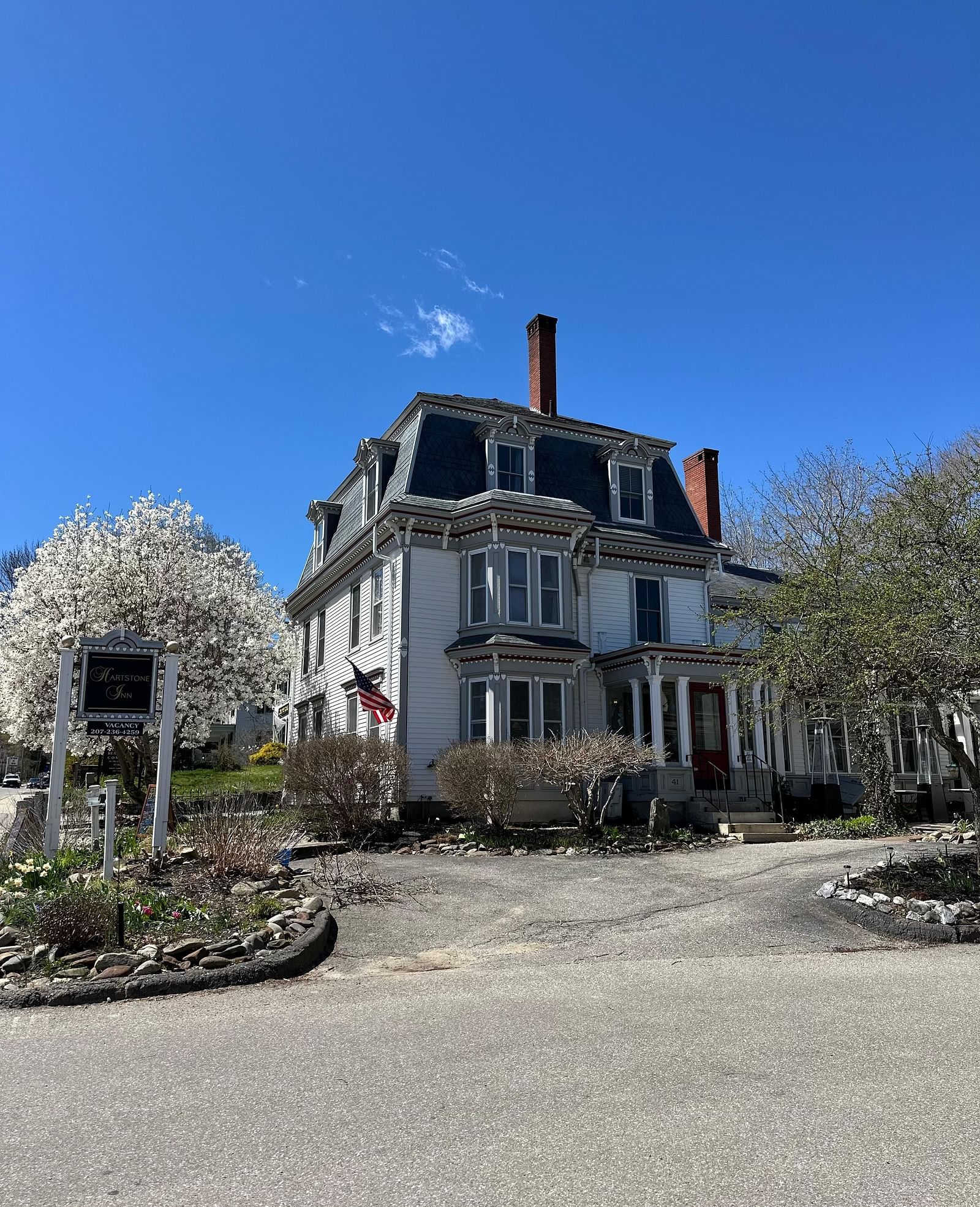 Hartstone Inn on a sunny day, showcasing its grand Victorian architecture, American flag on top, and blooming trees lining the driveway.