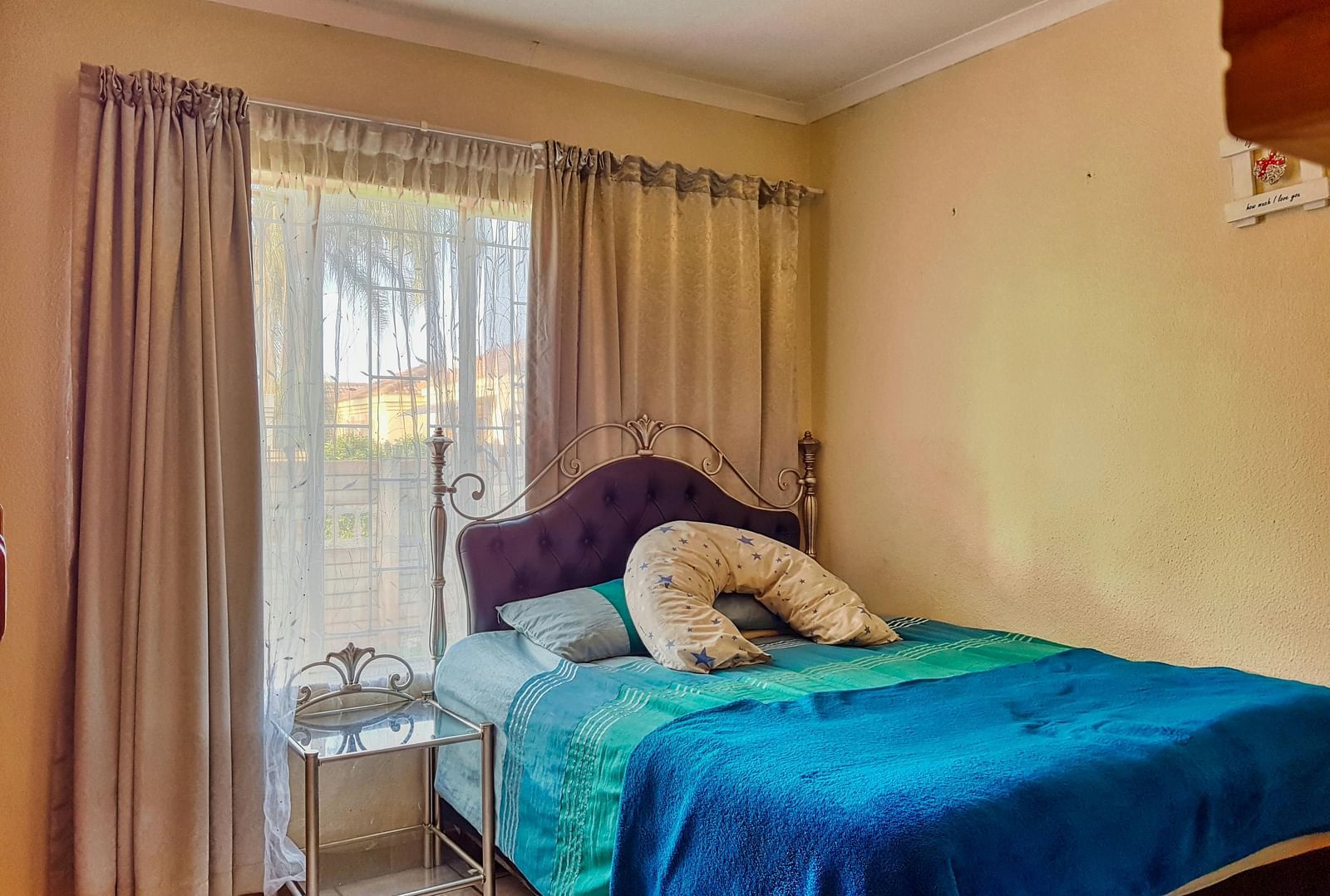 A bedroom featuring a double bed with blue sheets spread neatly over it with beige curtains covering the window behind it.