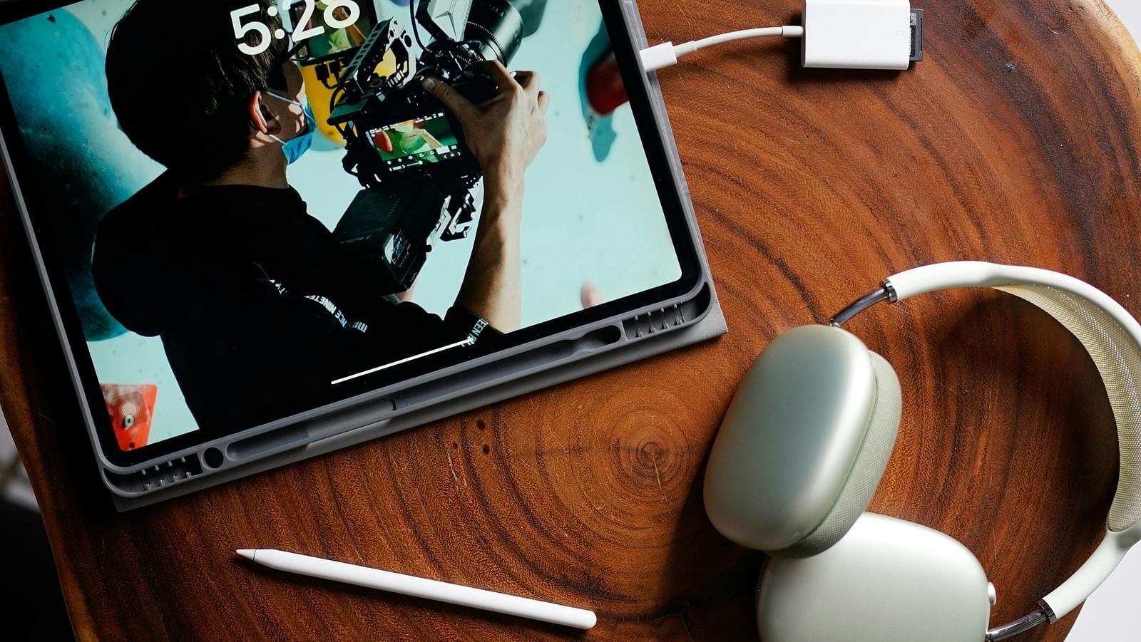 A tablet placed on a wooden surface with headphones, a stylus, and a charging adapter.
