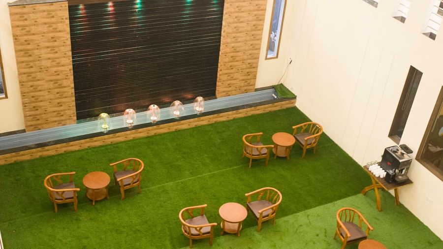 Overhead view of an indoor area with wooden chairs and tables on green artificial grass, facing a water wall | Daksh The Madhuvan Suites