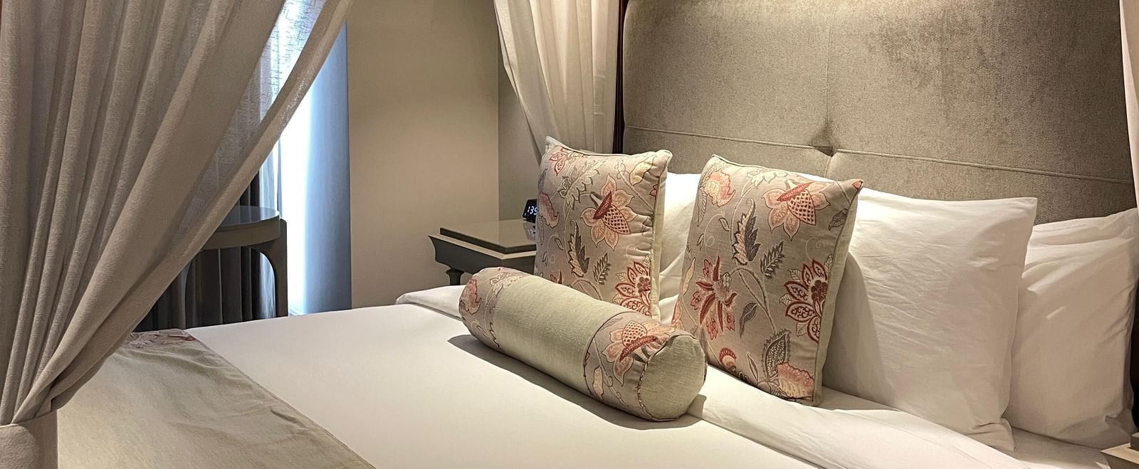 A side view of the four-poster bed in a Premium King Room at The Evren, Vagator. The bed is dressed with a bed runner and decorative pillows, and the white curtains are tied back, letting in natural light.