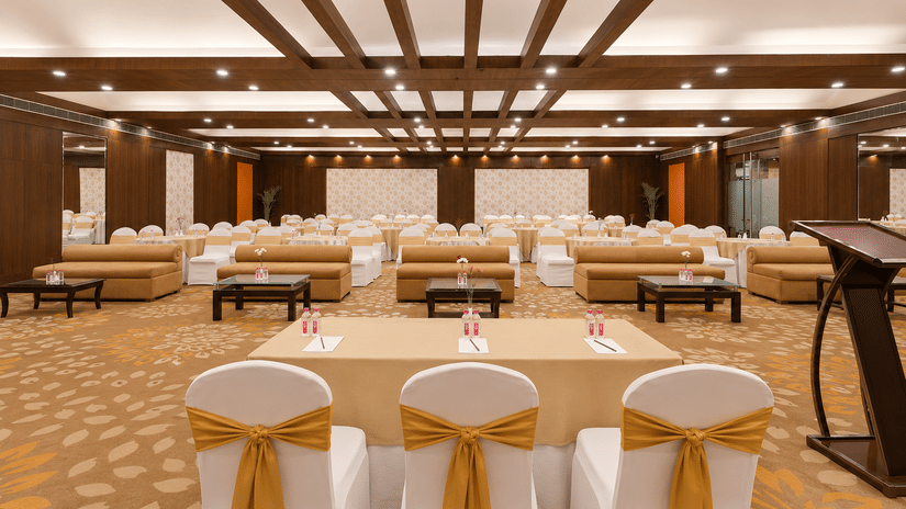 Elegant banquet hall with gold bow-tied chairs, wood ceiling beams, floral carpet, and conference setup.