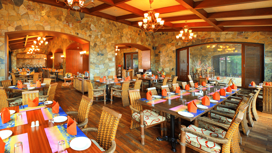 Amber restaurant at Ocean Spray Puducherry featuring wooden tables and chairs set, plates, cutlery, mat, and hand towels are placed on the table.