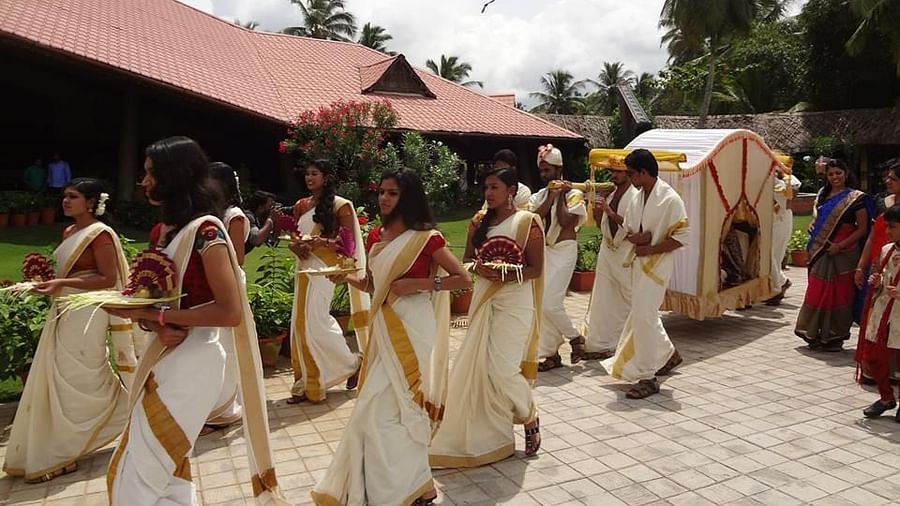 A group of people dressed in traditional attire, walking in procession with flower offerings, accompanied by a decorated palanquin in an outdoor setting