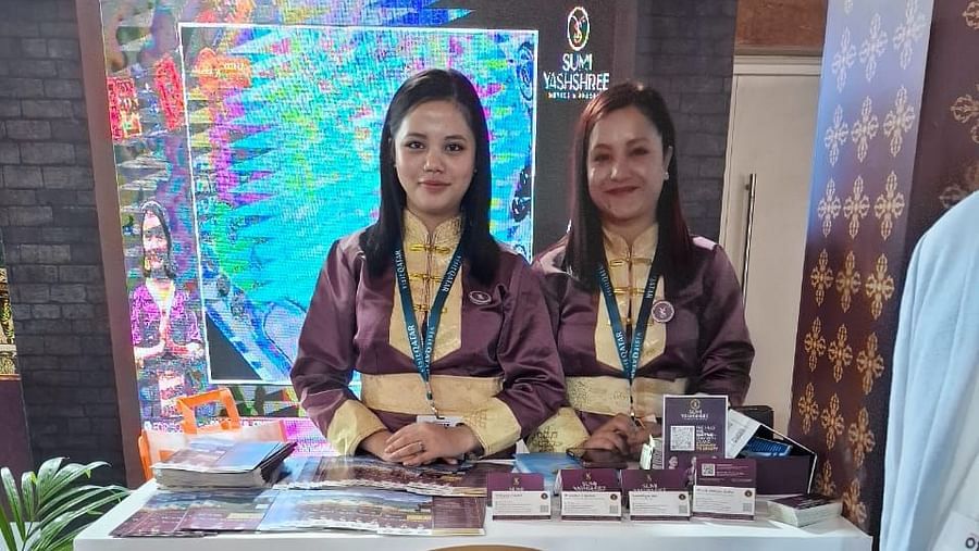 Two women in traditional uniforms at a YS branded counter with a digital screen backdrop.