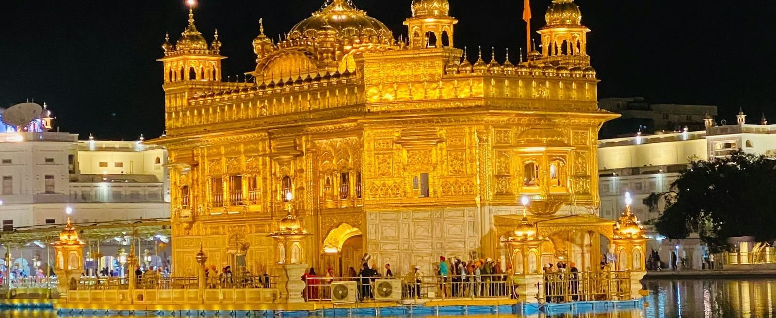 Exterior of a golden domed temple brightly illuminated at night, with a reflection in the water below.