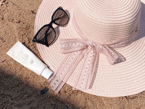 Sunscreen, a cap, and sunglasses displayed on sand during a sunny day.