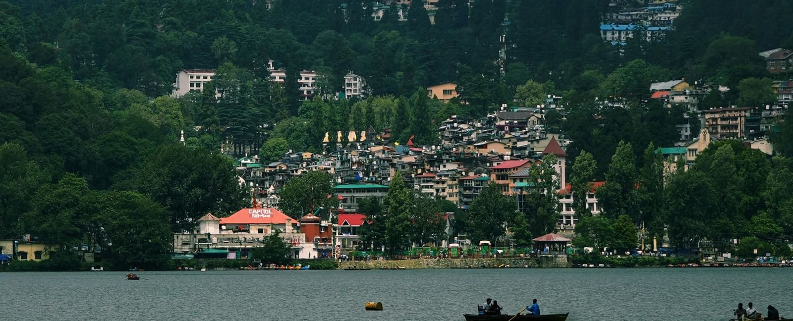 A close-up shot of a lake with small boats afloat, featuring a town settlement on the banks of the lake along with a cloudy sky in the background.