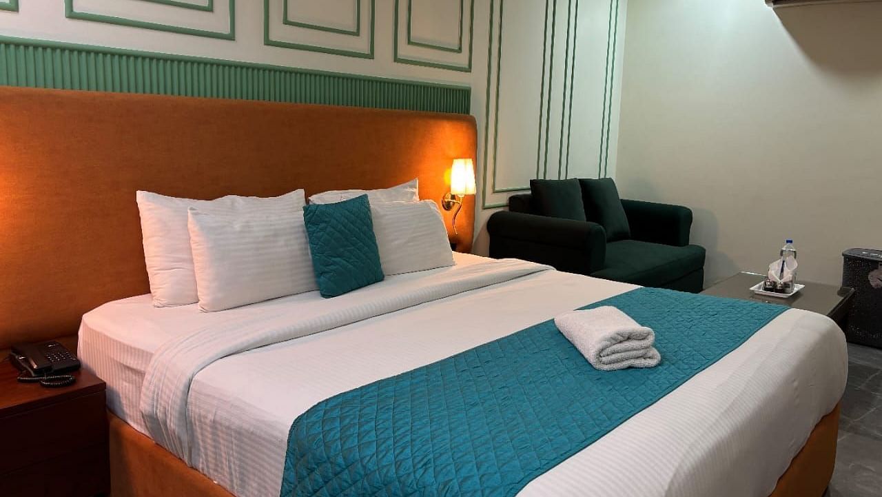 A well-lit room with a double bed dressed in white and blue, and a dark armchair - Asian Suites IFFCO Metro, Gurgaon