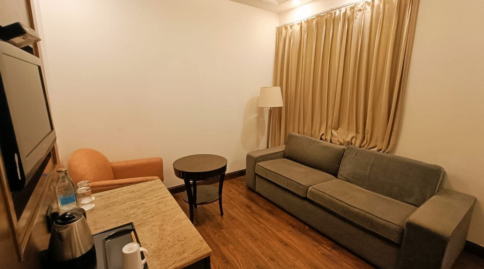 The suite's separate living room features a large sofa, wall-mounted television with a wooden table, lamp, and a single seating cushion at Inde Signature Tower, Gurgaon