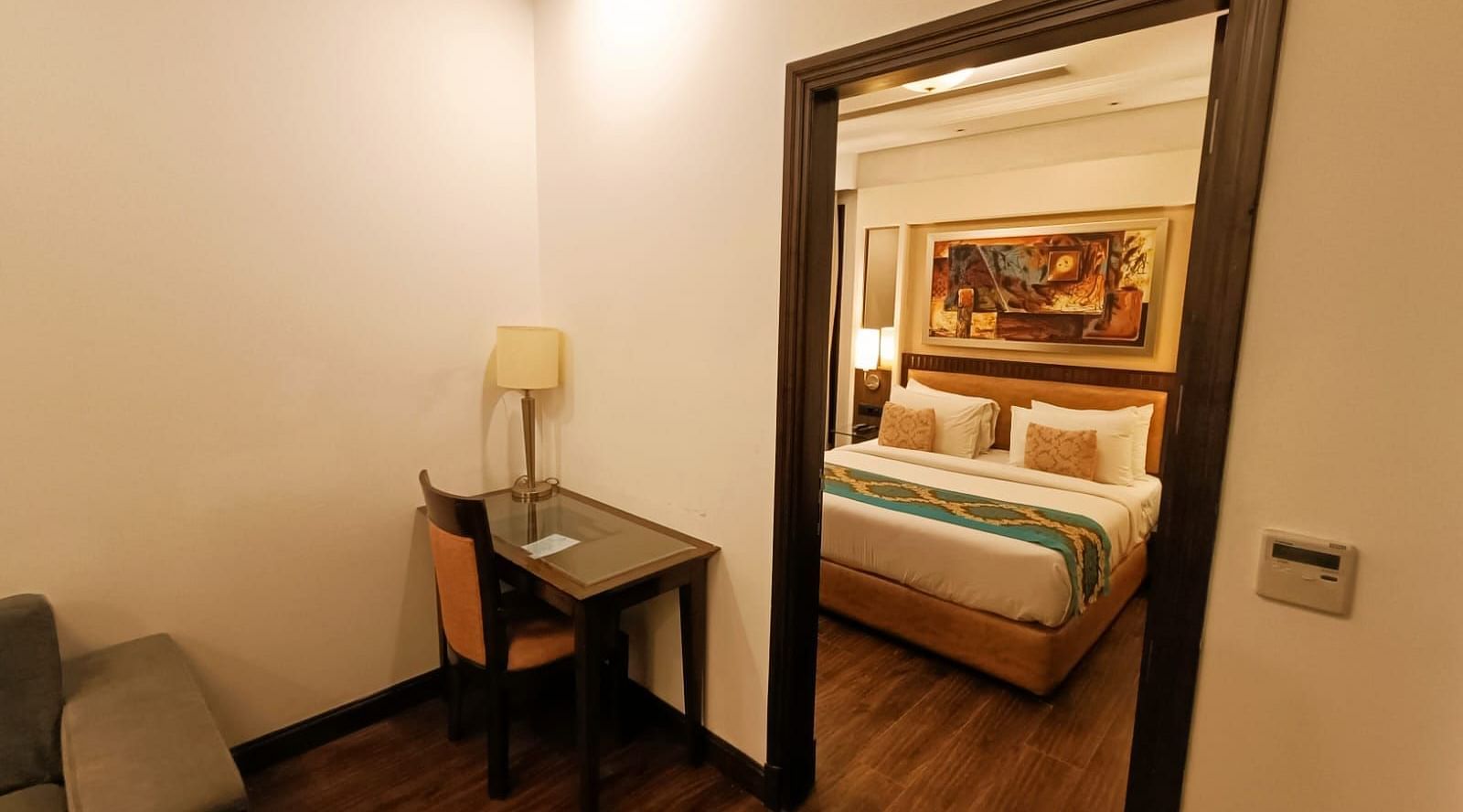Suite view from separate living area with a double bed neatly arranged with plush pillows, bedside tables each topped with a lamp, and a photo frame on the headboard at Inde Signature Tower, Gurgaon