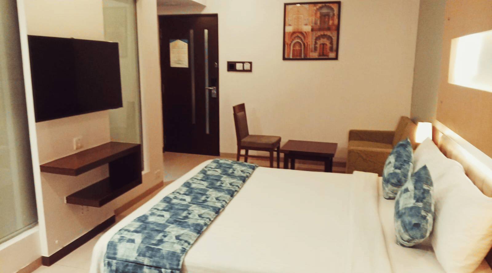 A hotel room layout showing a king size bed, a wall-mounted television, a wooden chair with a small side table, and an entry door at Hotel Mumbai House, Thane.