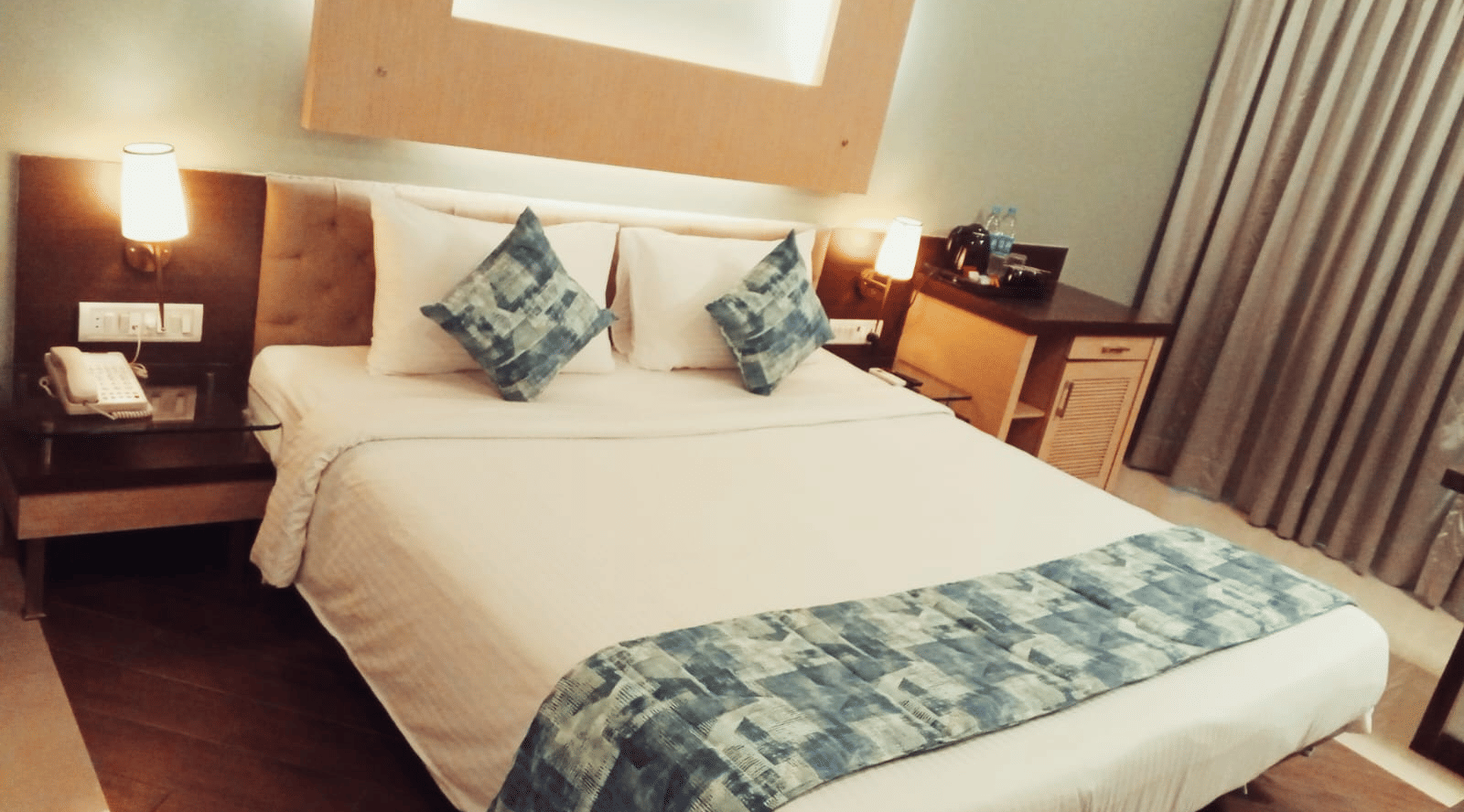 An angled view of a king size bed with a patterned bed runner, a small wooden bedside table holding a telephone, and an office chair at Hotel Mumbai House, Thane.