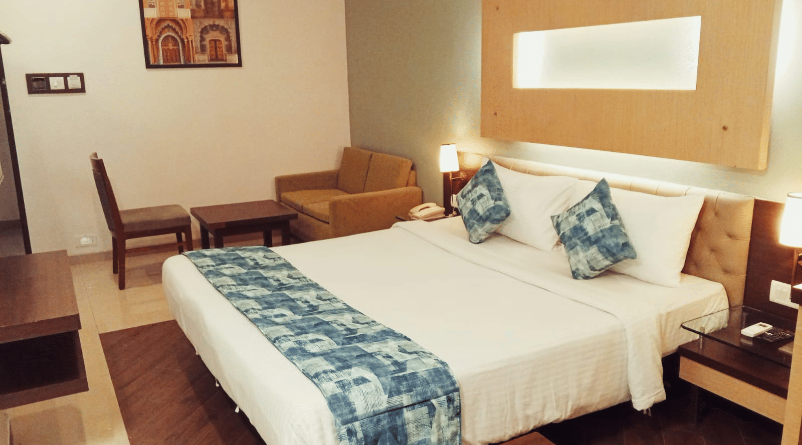 A bedroom layout showcasing a king size bed, a fabric sofa, a single chair, a wooden coffee table, and a framed wall painting at Hotel Mumbai House, Thane.