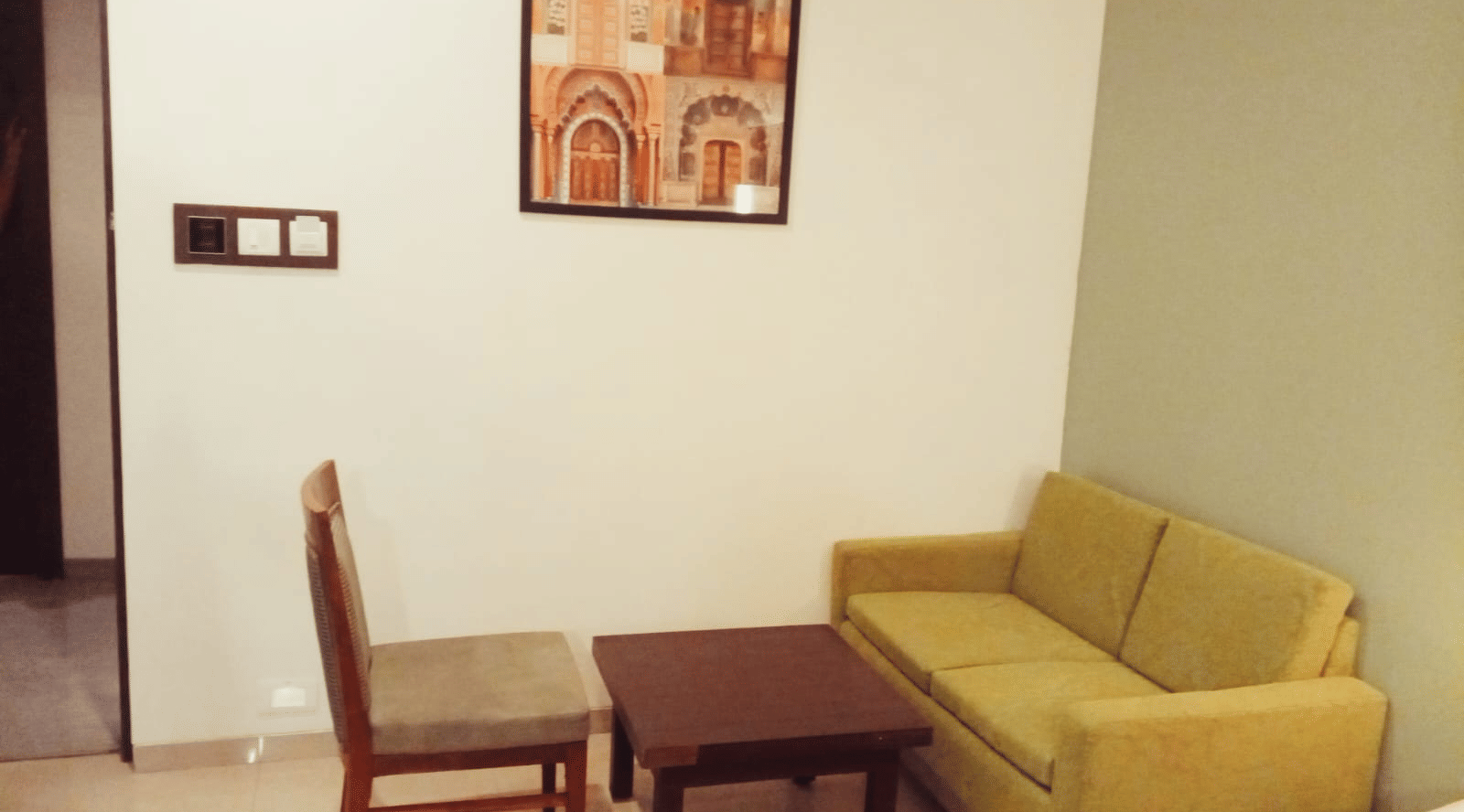 A seating area featuring a light fabric sofa, a rectangular wooden coffee table, and a single wooden chair against a plain wall at Hotel Mumbai House, Thane.