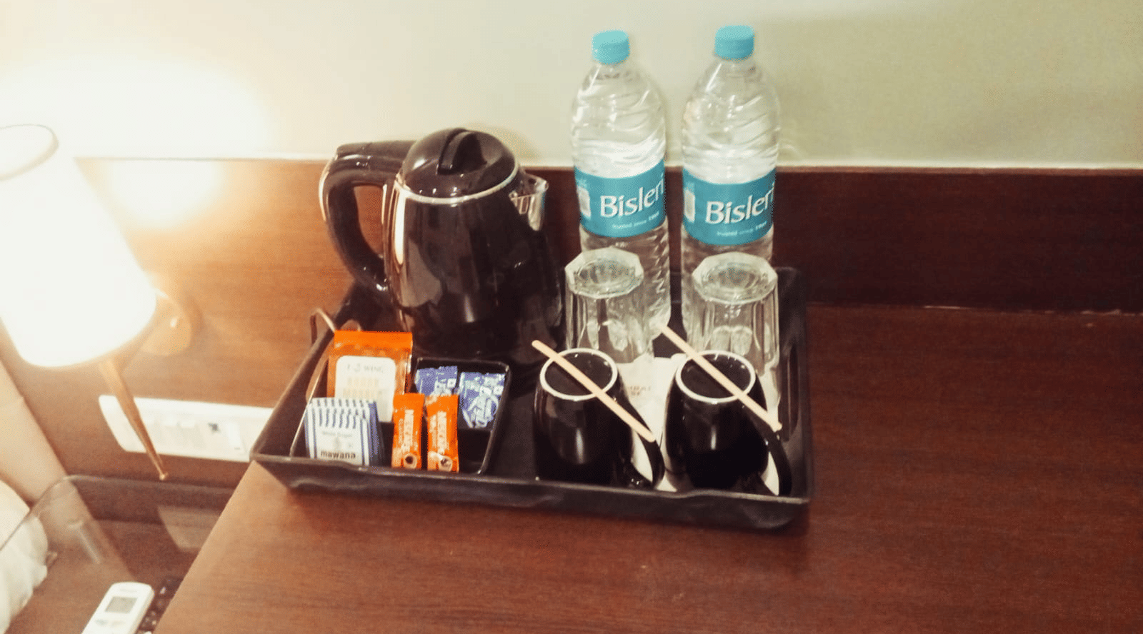 A close-up view of a black electric kettle, 2 plastic water bottles, 2 inverted glasses, and a selection of beverage sachets on a tray at Hotel Mumbai House, Thane.