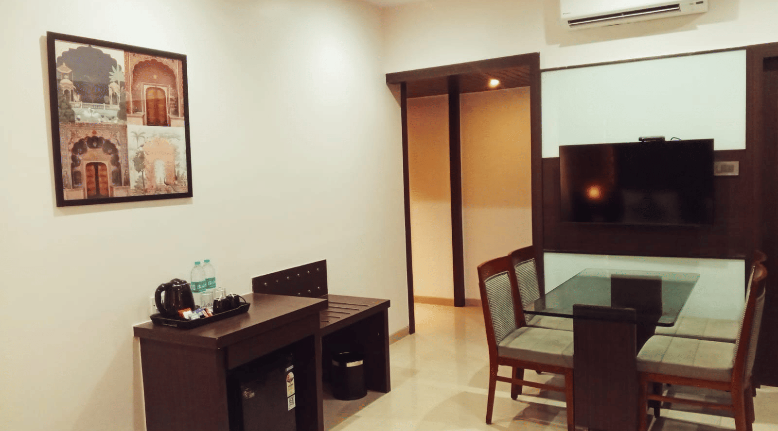 A bedroom at Hotel Mumbai House, Thane featuring a dining area with a glass top table, chairs, a dark wooden luggage desk, a framed picture, and a television.